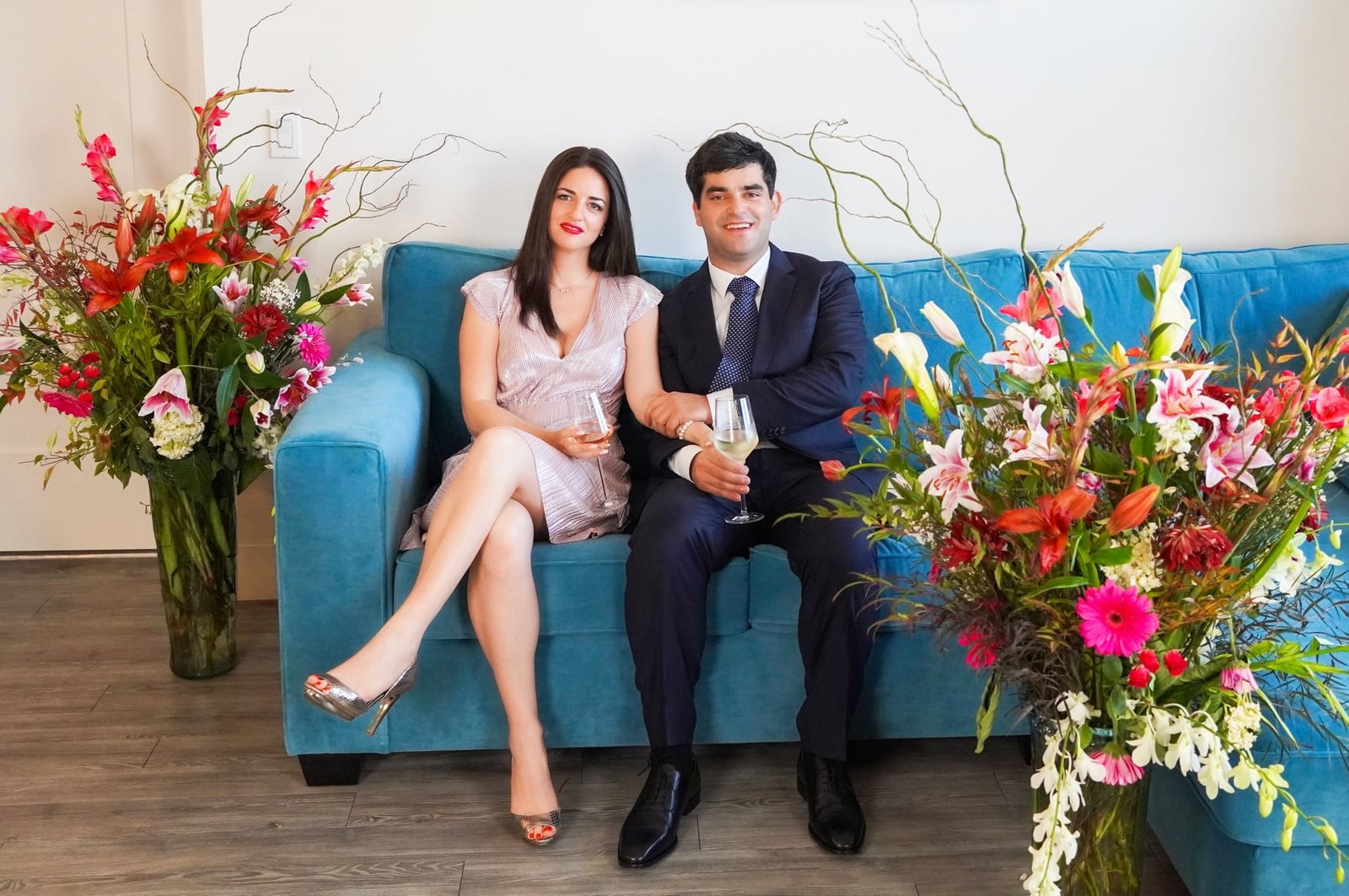 Polina and Dmitry seated on a teal sofa between large flower arrangements, holding glasses of champagne