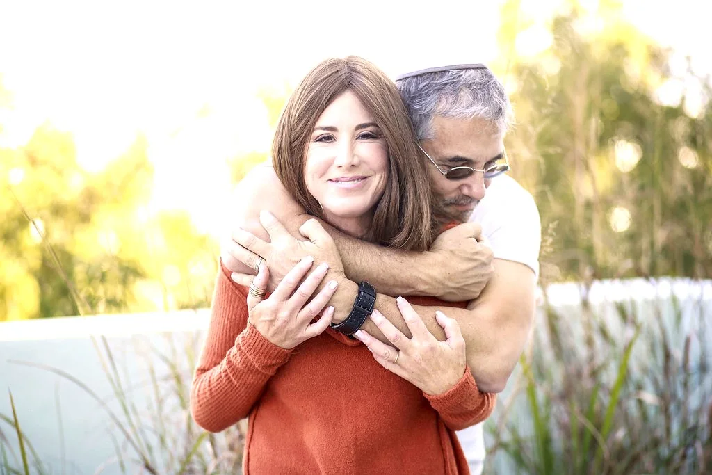 Michael embracing Bari from behind as they both smile outdoors