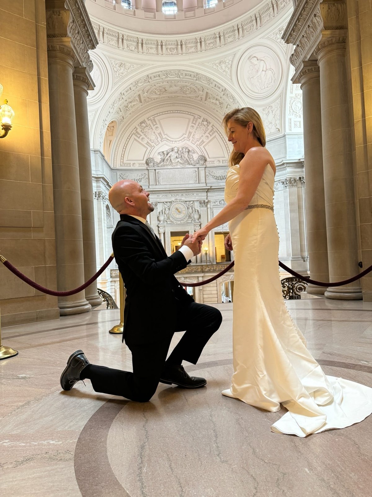Drew kneeling and holding Hayley's hand as she stands in her wedding gown beneath a domed rotunda